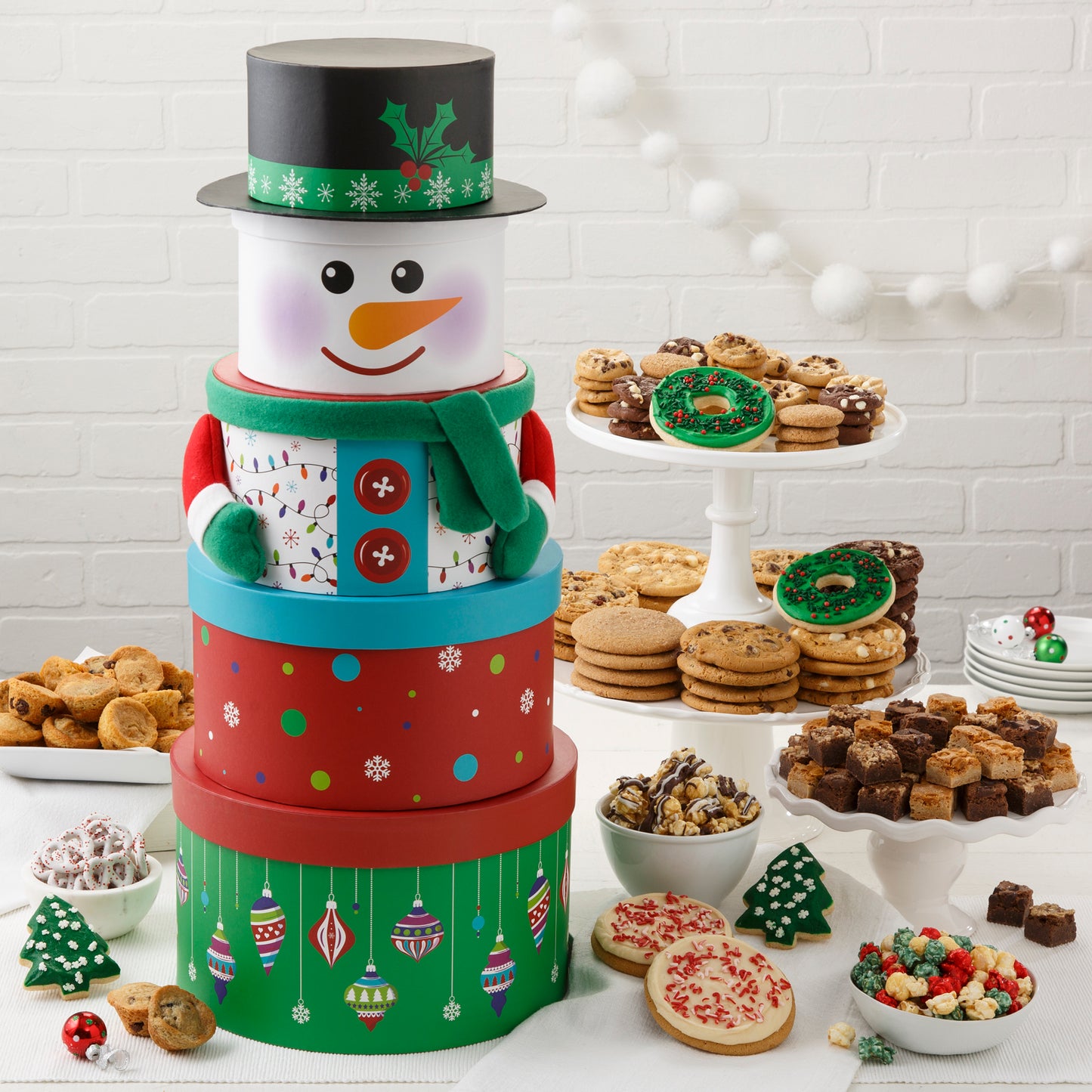 A five-tier snowman tower surrounded by an assortment of Nibblers®, original cookies, muffins, brownie bites, holiday popcorn, chocolate drizzled popcorn, two frosted wreath cookies with sprinkles, two frosted peppermint cookies with sprinkles, two frosted Christmas tree cookies with sprinkles, and yogurt-covered pretzels.