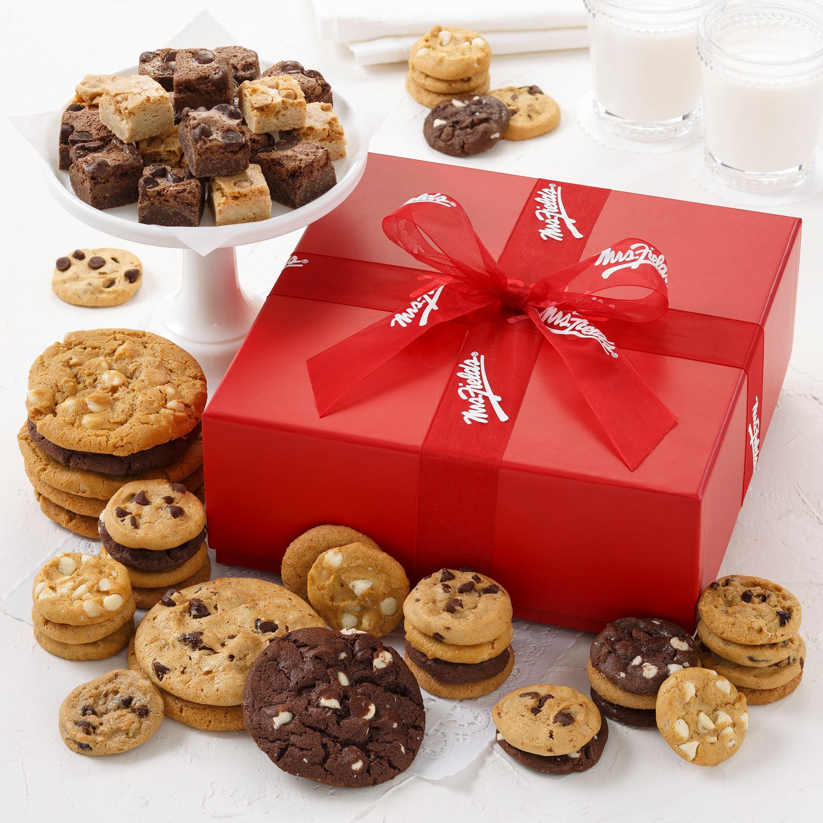 A red gift box tied with a red Mrs. Fields ribbon and surrounded by an assortment of Nibblers®, original cookies, and brownie bites.