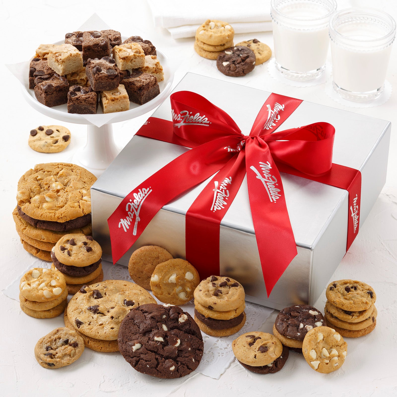 A silver gift box and tied with a red Mrs. Fields ribbon and surrounded by an assortment of Nibblers®, original cookies, and brownie bites