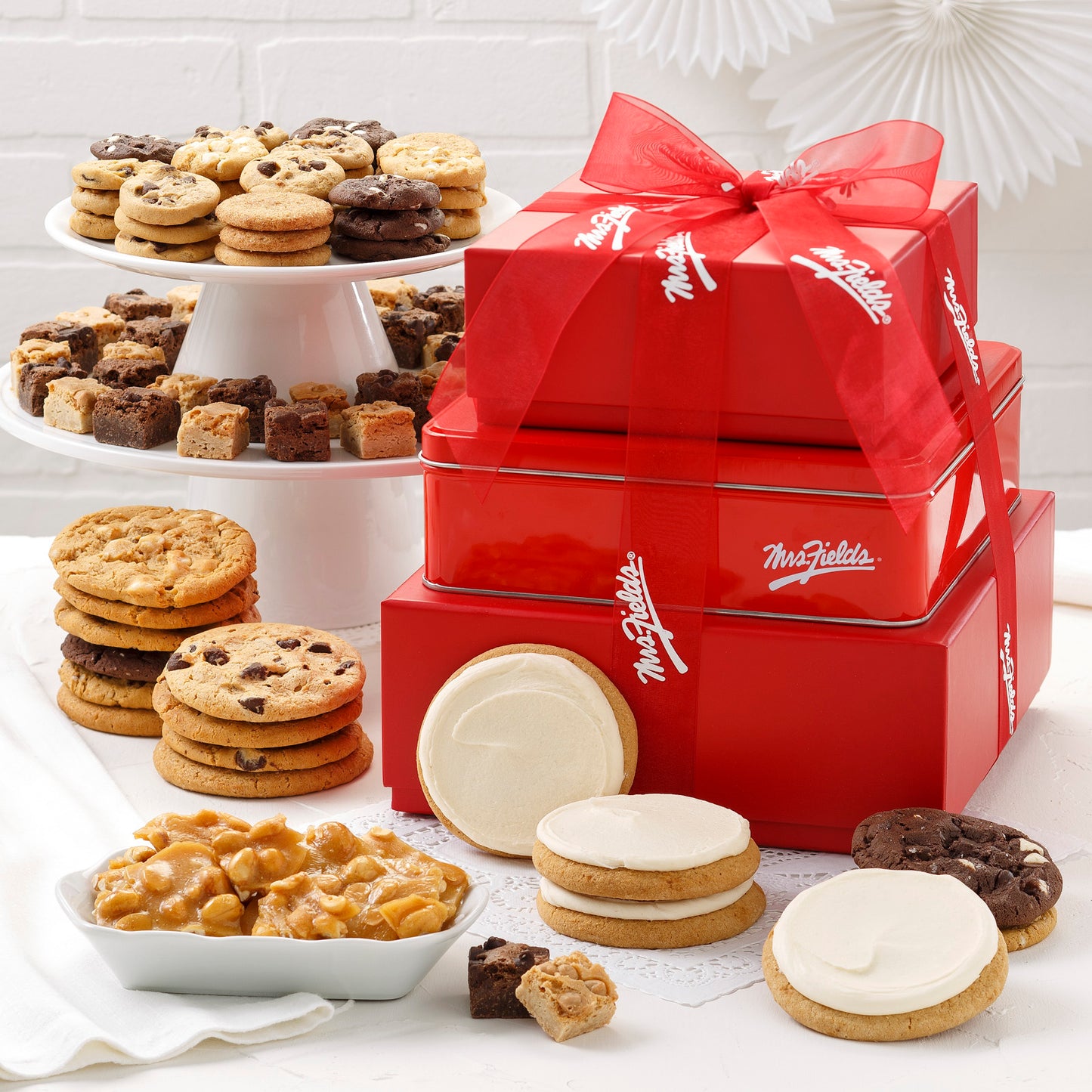 A three tier red tower tied with a Mrs. Fields ribbon and surrounded by an assortment of Nibblers®, original cookies, brownie bites, frosted cookies, and peanut brittle