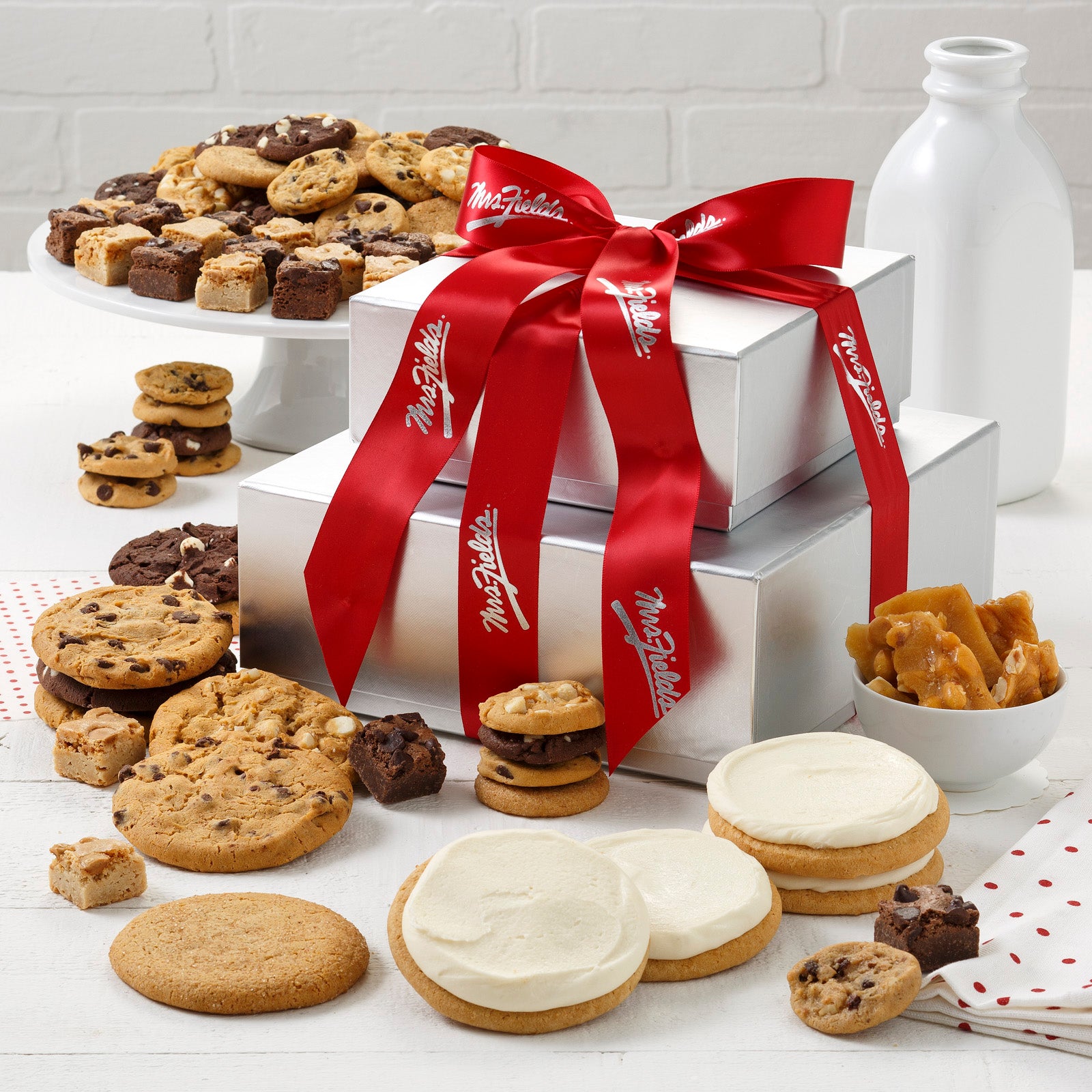 A two tier silver tower tied with a Mrs. Fields ribbon and surrounded by an assortment of Nibblers®, original cookies, brownie bites, frosted cookies, and peanut brittle