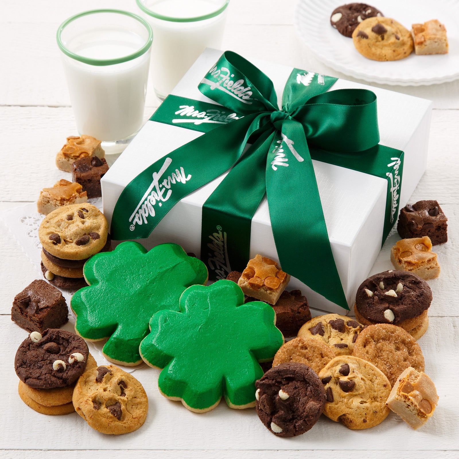 A white linen gift box tied with a green Mrs. Fields ribbon and surrounded by an assortment of nibblers, brownie bites, and two frosted shamrock cookies