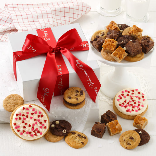 A white gift box wrapped with a Happy Valentine's Day red ribbon and surrounded with an assortment of nibblers, brownie bites, and two frosted round cookies with heart sprinkles.