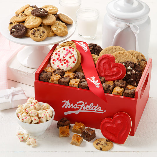 Red Mrs. Fields crate surrounded with original cookies, brownie bites, nibblers, red frosted heart cookies, a frosted round cookie with heart sprinkles, heart sprinkled popcorn and topped with a red Mrs. Fields ribbon. 