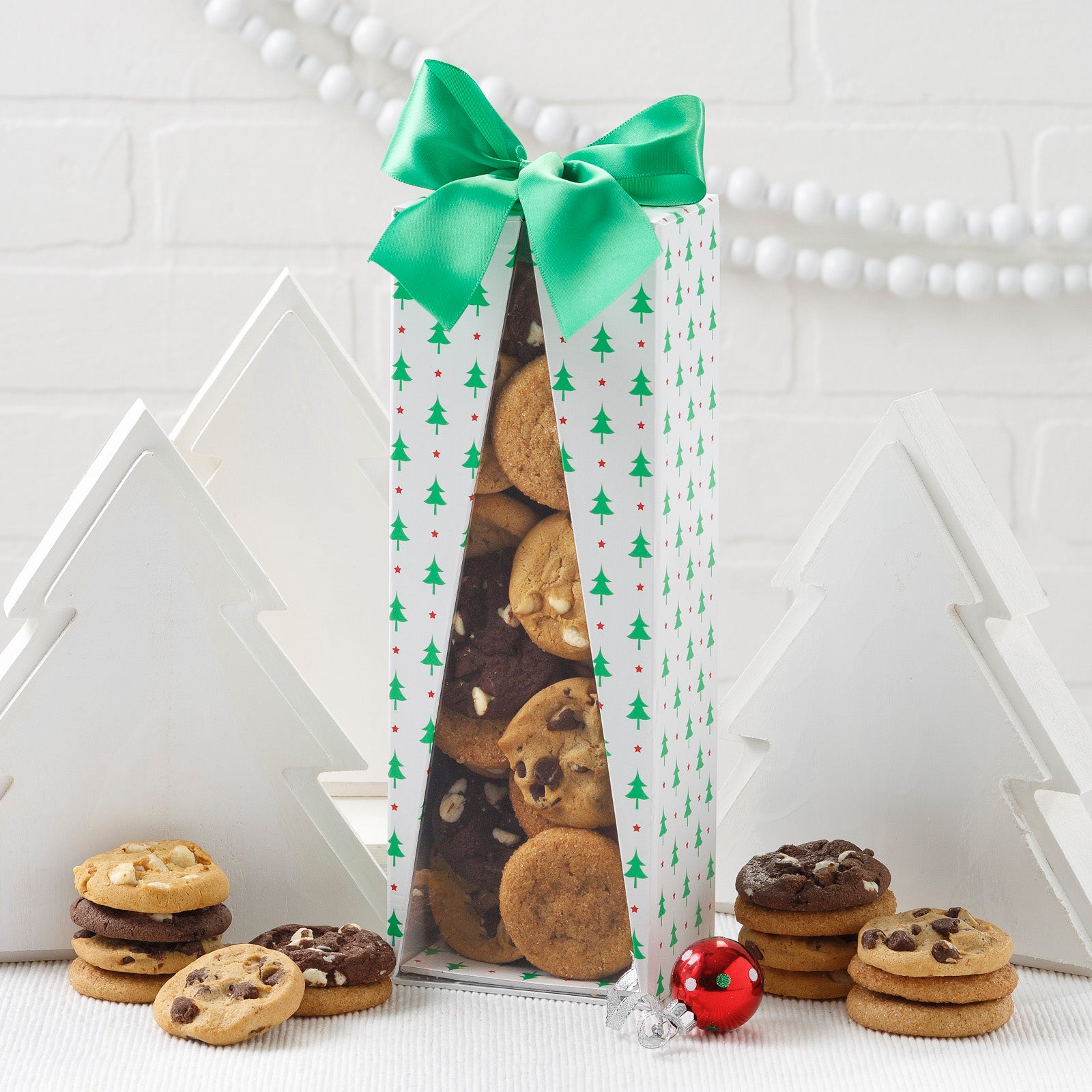 A holiday-themed gift box covered in trees with a green ribbon on top filled with an assortment of Nibblers® Bite-Sized Cookies.