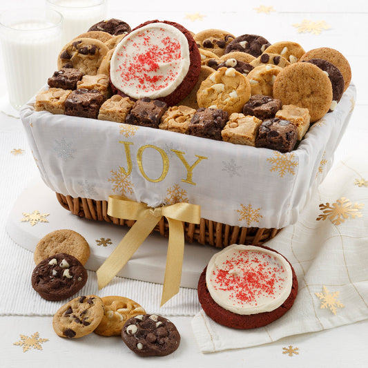 A basket decorated with a sentiment of Joy and covered with gold snowflakes and filled with an assortment of Nibblers®, brownie bites, one frosted red velvet cookies