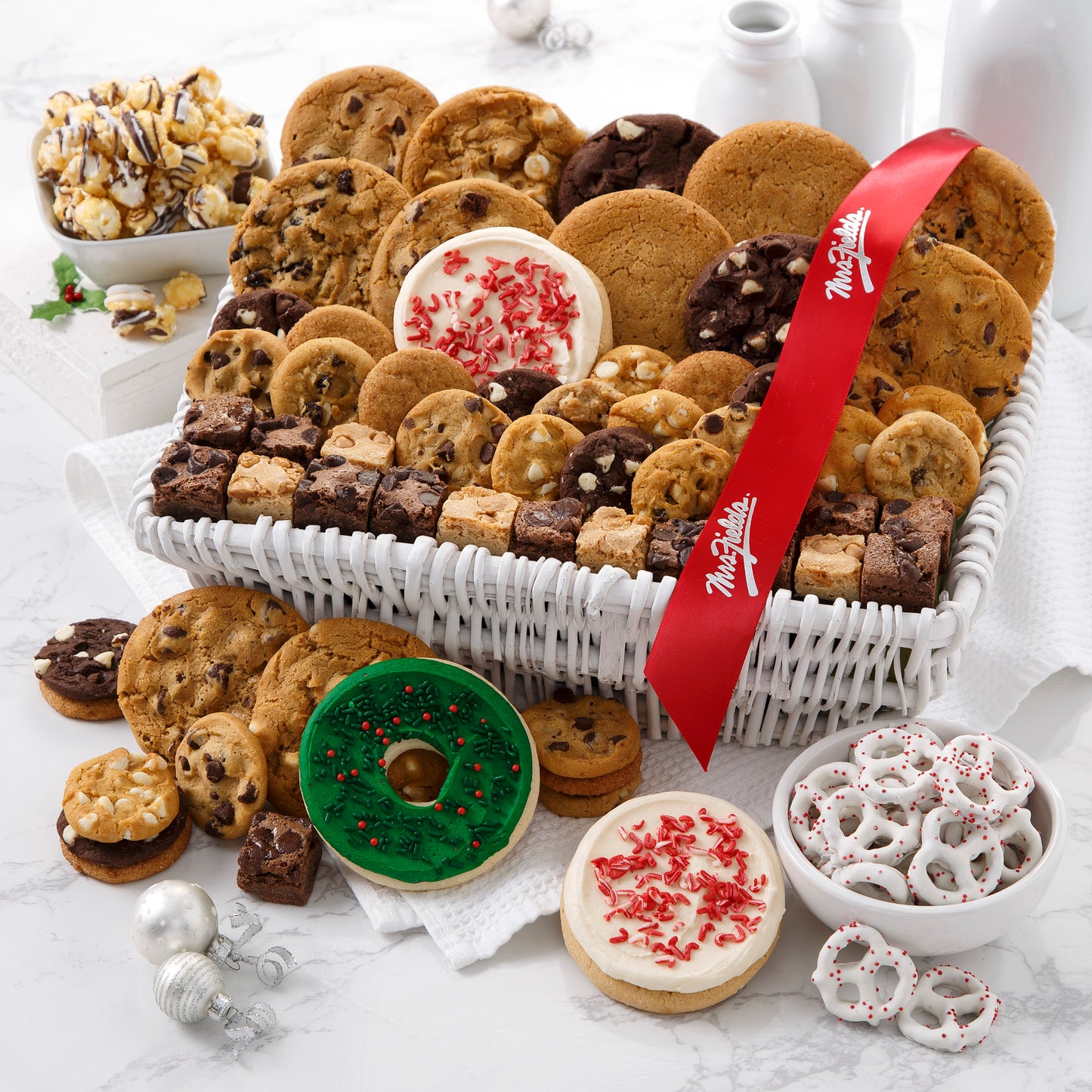 A white basket filled with an assortment of original cookies, Nibblers®, brownie bites, yogurt-covered pretzels, two frosted peppermint cookies, one frosted wreath cookies with sprinkles, and chocolate drizzled popcorn and decorated with a red Mrs. Fields' ribbon.