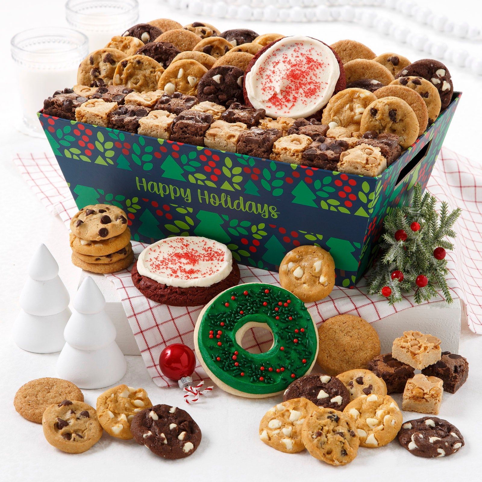 A Happy Holiday crate decorated with Christmas trees, holly and berries and filled with an assortment of Nibblers®, brownie bites, pretzels, two red frosted cookies, and one frosted wreath  cookie with sprinkles.