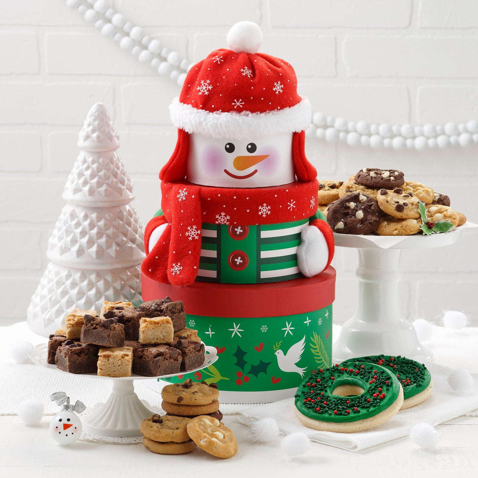 A green, red, and white snowman small tower that is surrounded by an assortment of Nibblers®, brownie bites, and two frosted wreath cookies with sprinkles.