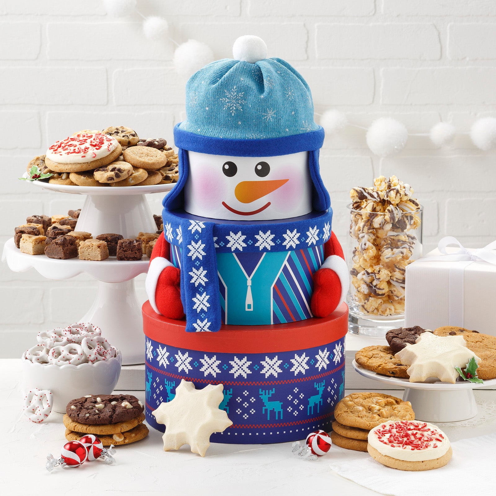 A blue, red, and white snowman tower surrounded by an assortment of Nibblers®, brownie bites, two frosted snowflakes, two frosted peppermint cookies with sprinkles, yogurt-covered pretzels, and chocolate drizzled popcorn.