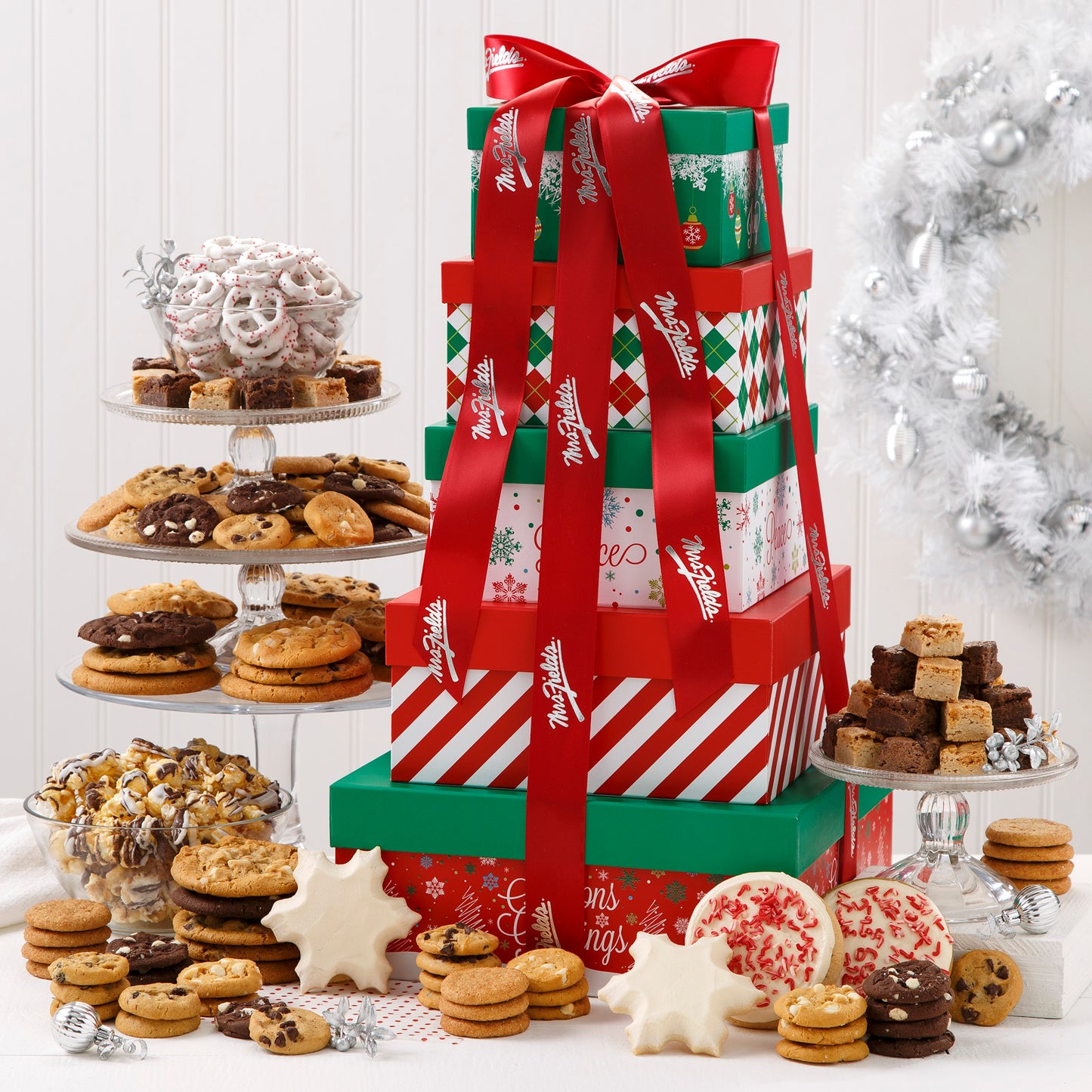 A five-tier red, green and white square tower tied at the top with a white Mrs. Fields ribbon and surrounded by an assortment of Nibblers®, original cookies, brownie bites, three frosted peppermint cookies with sprinkles, two frosted snowflakes, two peppermint sprinkled cookies, yogurt-covered pretzels, and chocolate drizzled popcorn.