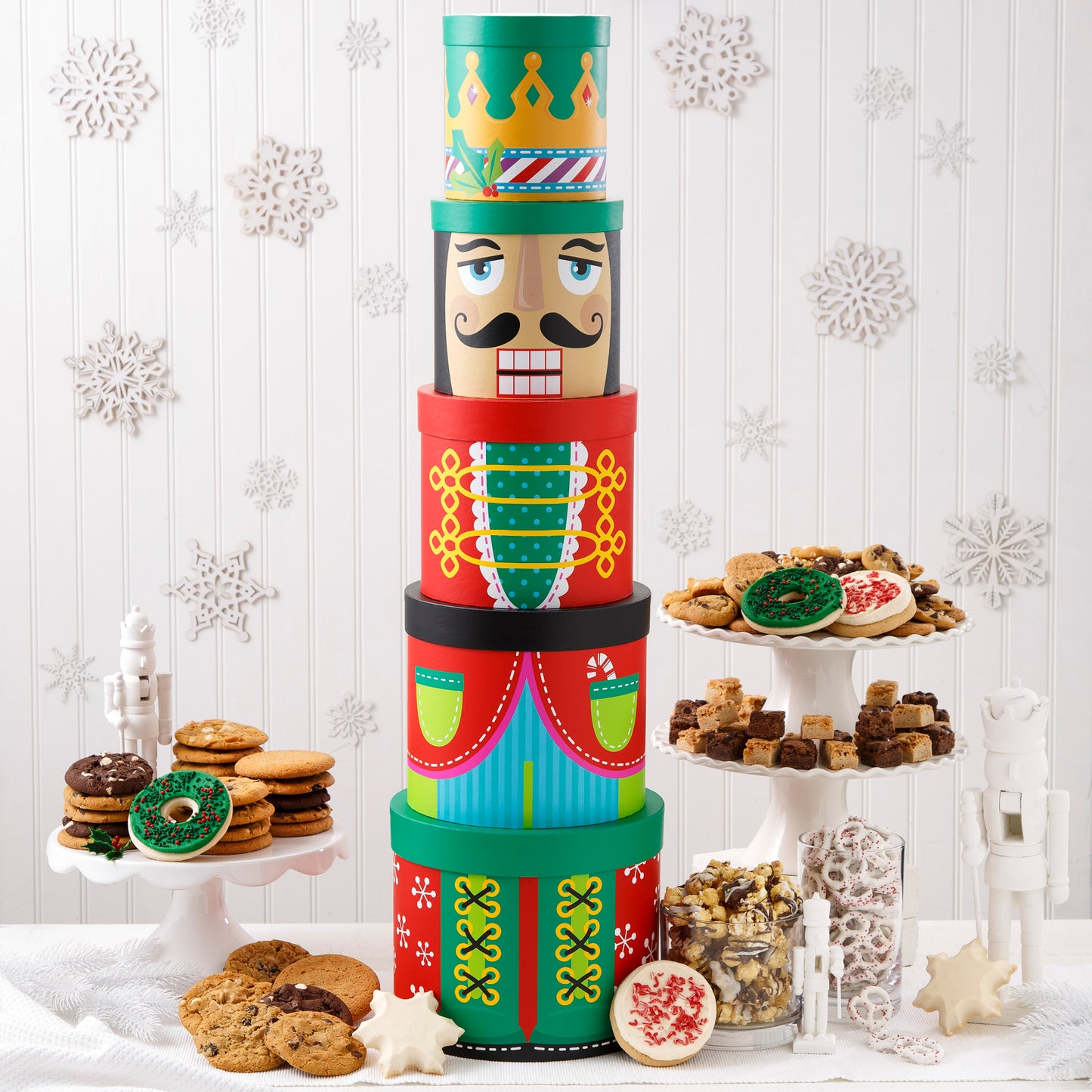 A five-tier nutcracker tower surrounded by an assortment of Nibblers®, original cookies, holiday popcorn, chocolate drizzled popcorn, yogurt-covered pretzels, two frosted wreath cookies with sprinkles, two snowflake cookies, and two frosted peppermint cookies with sprinkles.