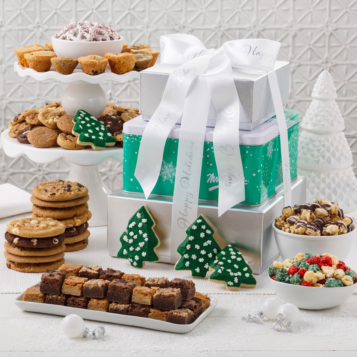 Green and silver three-tier tower tied with a white Mrs. Fields ribbon and surrounded by an assortment of Nibblers®, original cookies, brownie bites, popcorn, four frosted Christmas tree cookies with Sprinkles, muffins, and yogurt-covered pretzels.