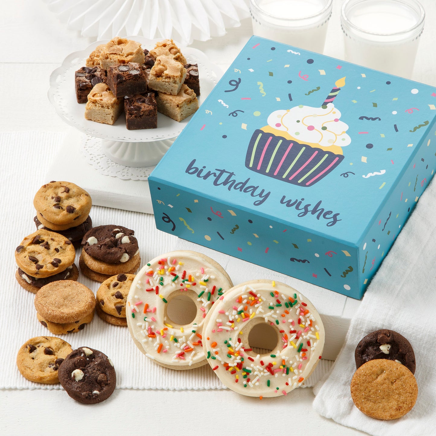 A blue Birthday wishes gift box surrounded by an assortment of nibblers, brownie bites, and two frosted white doughnut cookies