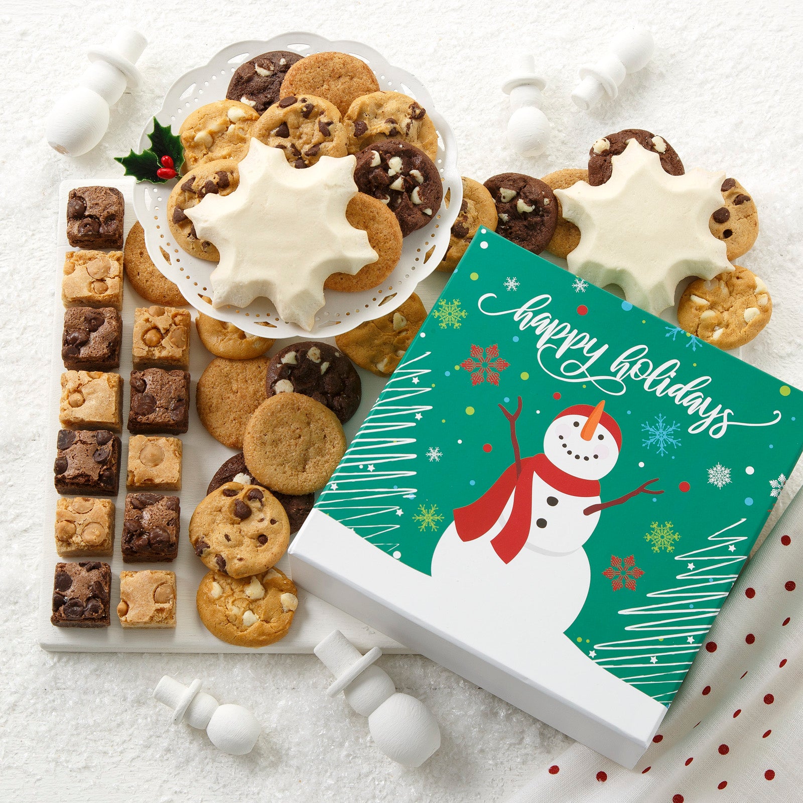 A Happy Holidays gift box decorated with a snowperson in a wonderland scene and surrounded by an assortment of Nibblers®, brownie bites and two frosted snowflake cookies 