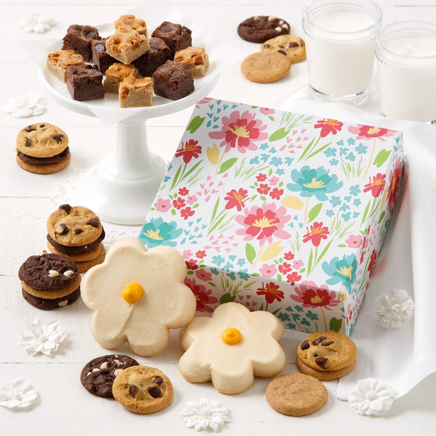 A white gift box decorated with flowers and surrounded by an assortment of nibblers, brownie bites, and two frosted daisy cookies