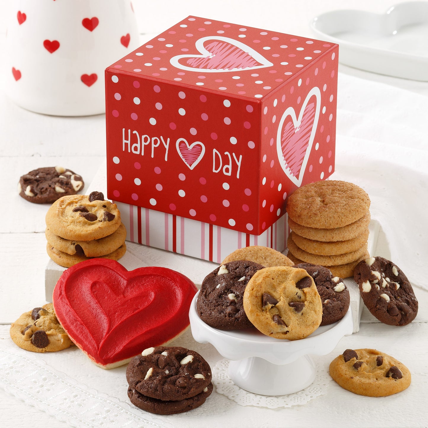 A red mini Happy Valentine's Day gift box decorated with dots and stripes and surrounded by an assortment of Nibblers and one red frosted heart cookie.