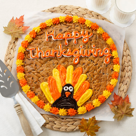 A cookie cake decorated with yellow and orange frosting with a festive turkey in frosting as decoration.